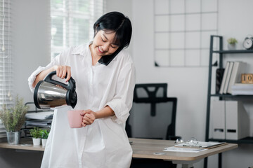 A young woman in a white shirt pours coffee into a mug while talking on the phone in a modern office with natural light and minimalist decor.