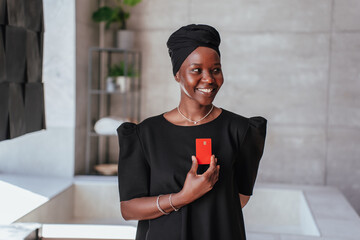 Black woman in a black dress and headwrap holds a red card and smiles warmly in a modern, minimalist indoor setting