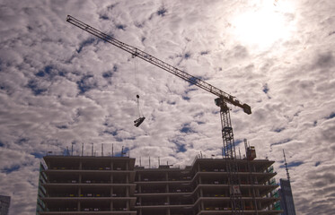 Construction crane lifts metal structures, in the background a cloudy summer sky and the sun breaking through the clouds, real estate market concept.