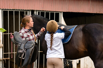 Girl adjusting saddle pad on horse while instructor observes.
