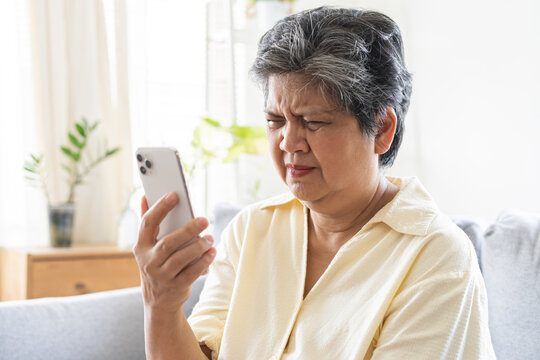 Presbyopia, Hyperopia mature, aged asian woman having vision problem trying to stare text on smart mobile phone screen on sofa at home, eye disease of elderly. Poor eyesight farsightedness concept.