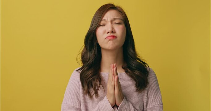 Young Asian woman shows signs of begging for mercy in need of something. Isolated on yellow background in studio.