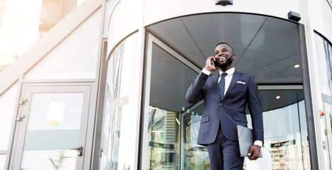 Business Workspaces For Rent. African American Businessman Talking On Cellpone Against Entrance To Modern Coworking Office Building. Low-Angle