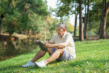 asian senior man sitting on grasses in the nature park,reading a book,mature male relaxing in the morning sunshine