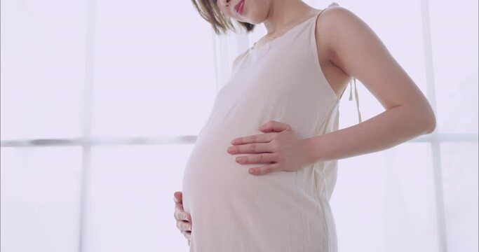 Happy Asian pregnant woman lovingly touching her unborn baby while standing by the window at home.