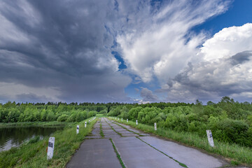 A winding paved road leads through a lush green forest with a dramatic sky above, featuring a mix of dark storm clouds and bright white clouds.