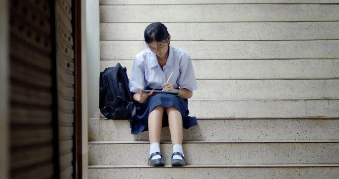 Thai or Asian high school girls in cute white school uniforms, using tablets and pens for practicing reading and revising knowledge, sitting on school stairs in the quiet evening after school.