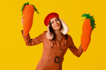 Stylish young woman in bunny ears holding toy carrots on yellow background. Easter celebration