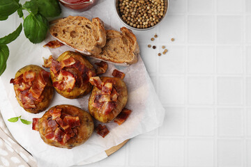 Delicious baked potatoes with bacon, bread and peppercorns on white tiled table, flat lay. Space for text