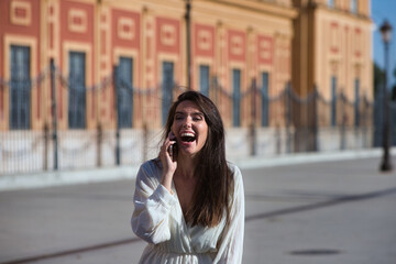 Fototapeta premium Young and beautiful Latin woman talking on mobile phone is having fun and is happy and smiling loudly. The woman is on the street and walks through the city. Concept communications.