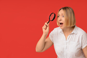 Shocked adult woman holding magnifying glass on red background