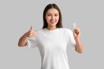 Young Asian woman with floss toothpicks showing thumb-up on grey background