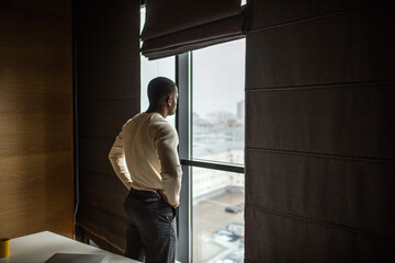 Serious dark skinned businessman, feeling worried about new business project, dressed in knitted white pullover, spends free time at office, looking at cityscape through pamoramic window.