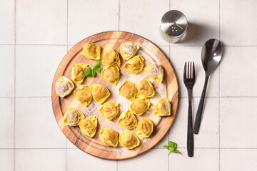 Wooden board with raw dumplings and basil on white tile background
