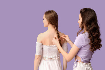 Beautiful young woman braiding hair for sister on lilac background