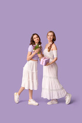 Beautiful young sisters with gift box and tulips on lilac background