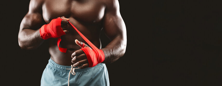 A muscular man with bare chest is wrapping his hands with red tape in preparation for a boxing or martial arts workout.