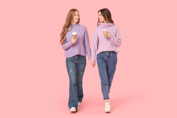 Beautiful young sisters with paper cups of coffee holding hands and walking on pink background