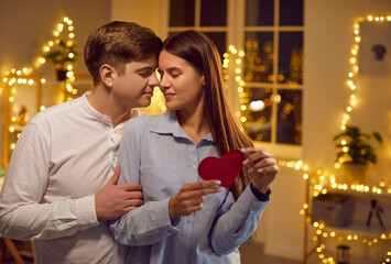 Portrait of cheerful smiling young couple embracing each other with tenderness standing with closed eyes at home with bokeh background. Happy man hugging his loving wife. Valentines day concept.
