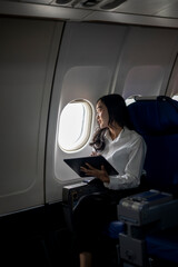 A woman is sitting in an airplane seat with a book and tablet in front of her