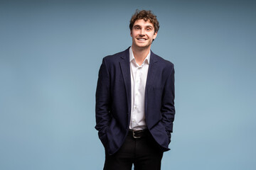 Confident young businessman smiling with hands in pockets on blue background looking at camera