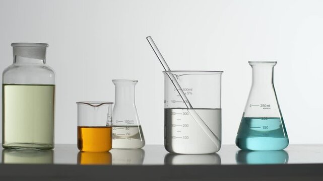 Close-up of an erlenmeyer flask containing a blue liquid placed on a shelf by a scientist. The camera pans across a series of lab glassware with colorful liquids, front angle video for advertising