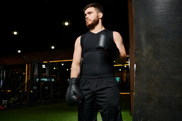A bearded man in a black tank top throws punches at a hanging punching bag in a gym setting.