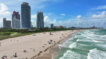 People enjoying Miami Beach with downtown skyline in background. Aerial rising shot over ocean waves.