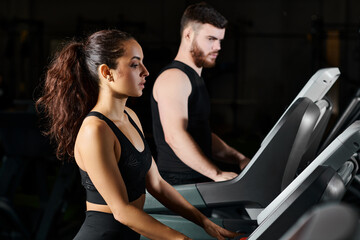Fototapeta premium A male trainer and a brunette sportswoman are exerting themselves while running on a treadmill in a gym.