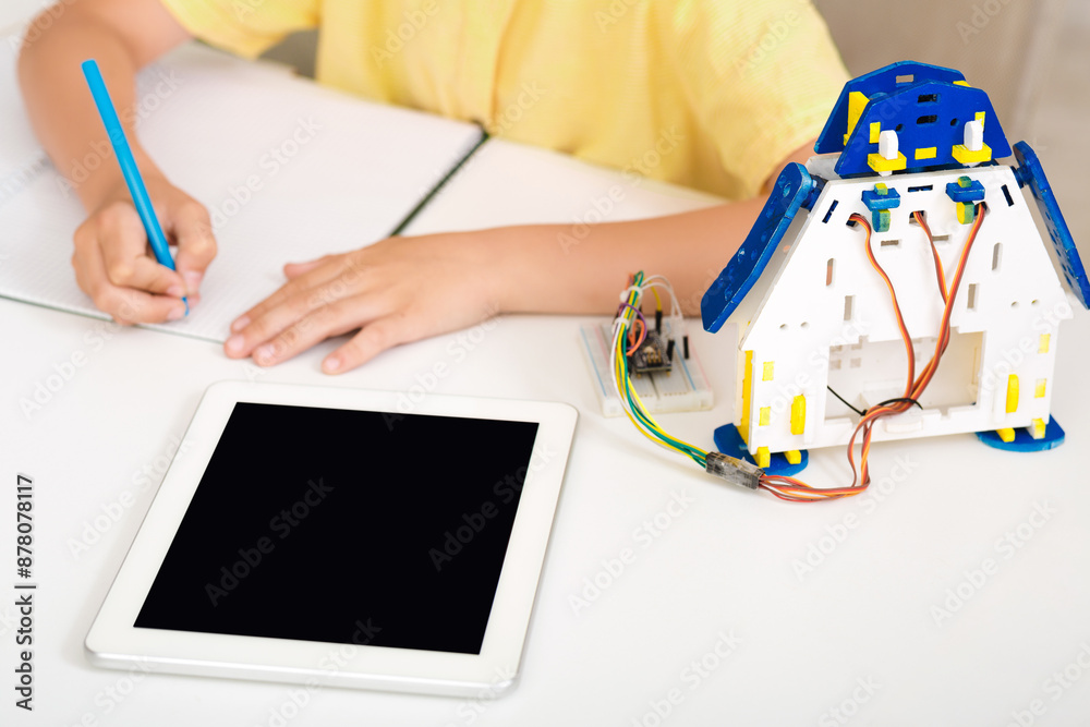 Poster A child is sitting at a desk, learning about robotics and coding. They are using a tablet computer and a robot kit. The childs hand is shown writing on a piece of paper