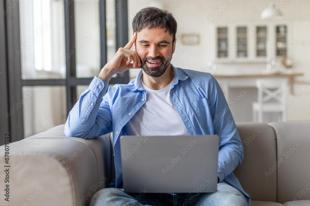 Aufkleber a man is sitting on a light gray couch while using a silver laptop. he is wearing a light blue butto