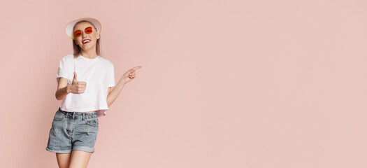Young woman dressed in a white t-shirt, denim shorts, and a white hat is smiling brightly while posing with a thumbs-up gesture and pointing to the right. She is standing in front of pink background.