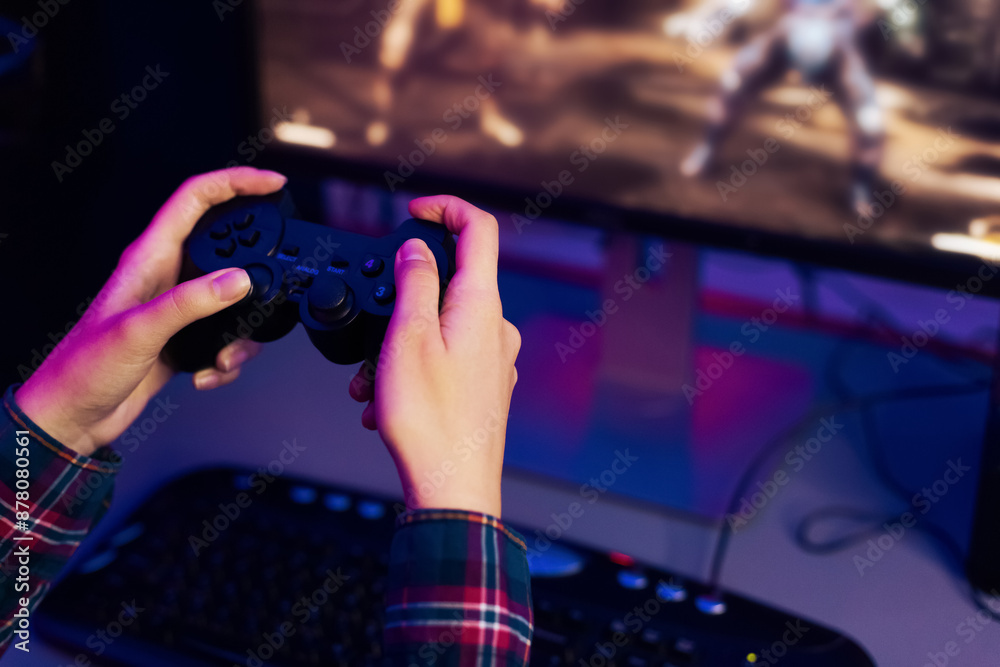 Aufkleber This is a photo of a persons hands holding a video game controller, with a computer monitor in the background displaying an action game. The person is immersed in the game, focused on the screen.