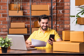 Indian asian Man Using Laptop and Smartphone to Manage and Ship Online Orders from Warehouse