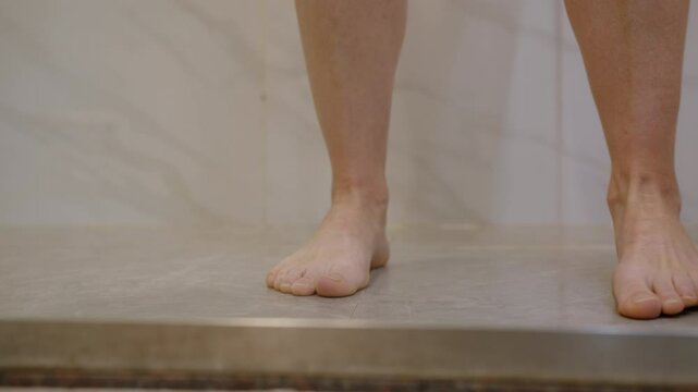 Woman walks barefoot into shower cabin and closes glass door in bathroom.
