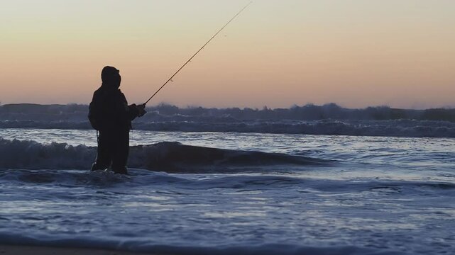 Un p&ecirc;cheur, les jambes dans l'eau, remonte sa ligne au bord de l'oc&eacute;an atlantique, en fin de soir&eacute;e, pendant l'heure bleue