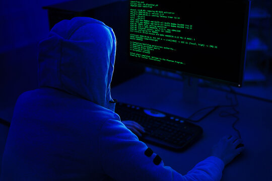 A person wearing a hooded sweatshirt sits in front of a computer with a dark background. Computer screen displays green code, indicating that the person is working on a programming or technical task. - Powered by Adobe