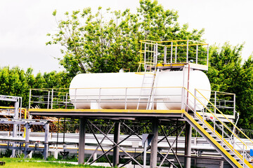 A white storage tank sits on an elevated platform, supported by metal beams. A staircase with a...