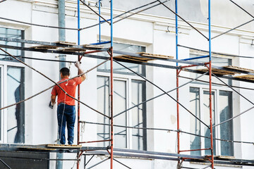 A construction worker paints the exterior of a building while standing on a scaffolding