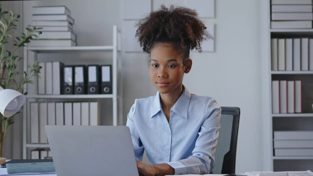 Young woman is focused on her work, typing on a laptop and taking notes in a modern office. Shelves and plants create a professional work environment