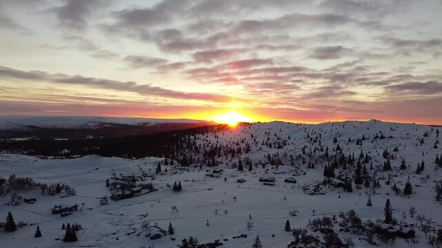 Breathtaking aerial view of a sunset casting a warm glow over the snowy landscape of Trysil, Norway, with scattered trees and cabins.