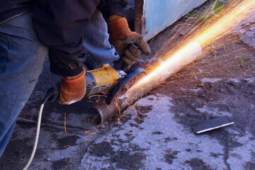 A worker wearing protective gloves cuts a metal pipe with a grinder, creating a shower of sparks. The worker is kneeling on the ground, holding the grinder with both hands.