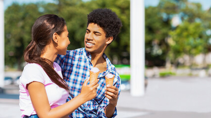 Obraz premium A teen black couple stand close together and smile as they each hold a waffle cone ice cream. They are standing outside on a sunny day, copy space