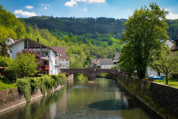Fototapeta premium Ein Fluss, der eingerahmt von Mauern, Bäumen und Häusern auf eine alte Brücke zuläuft, mit Waldhang im Hintergrund
