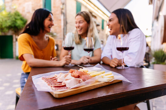 Fototapeta Three happy diverse women having fun together enjoying red wine at restaurant. Focus on wooden board with cheese and ham.