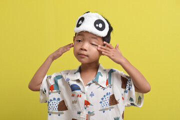 Portrait of Asian child wearing pajamas and sleep mask rubbing his eyes isolated on yellow background. Child wants to sleep. Healthy sleep concept. Copy space