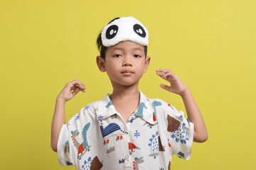 Portrait of Asian child stretching arms, wearing pajamas and sleep mask isolated on yellow background. Child wants to sleep. Healthy sleep concept. Copy space