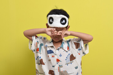 Portrait of sleepy Asian child rubbing his eyes isolated on yellow background. Child wearing pajamas and sleep mask. Healthy sleep concept. Copy space