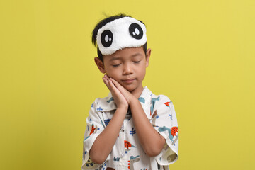 Portrait of cute Asian child wearing pajamas and sleep mask putting his hand under his cheek isolated on yellow background. Child wants to sleep. Healthy sleep concept