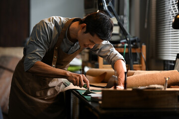 Small business craftsman in apron carefully cutting piece of leather in his workshop